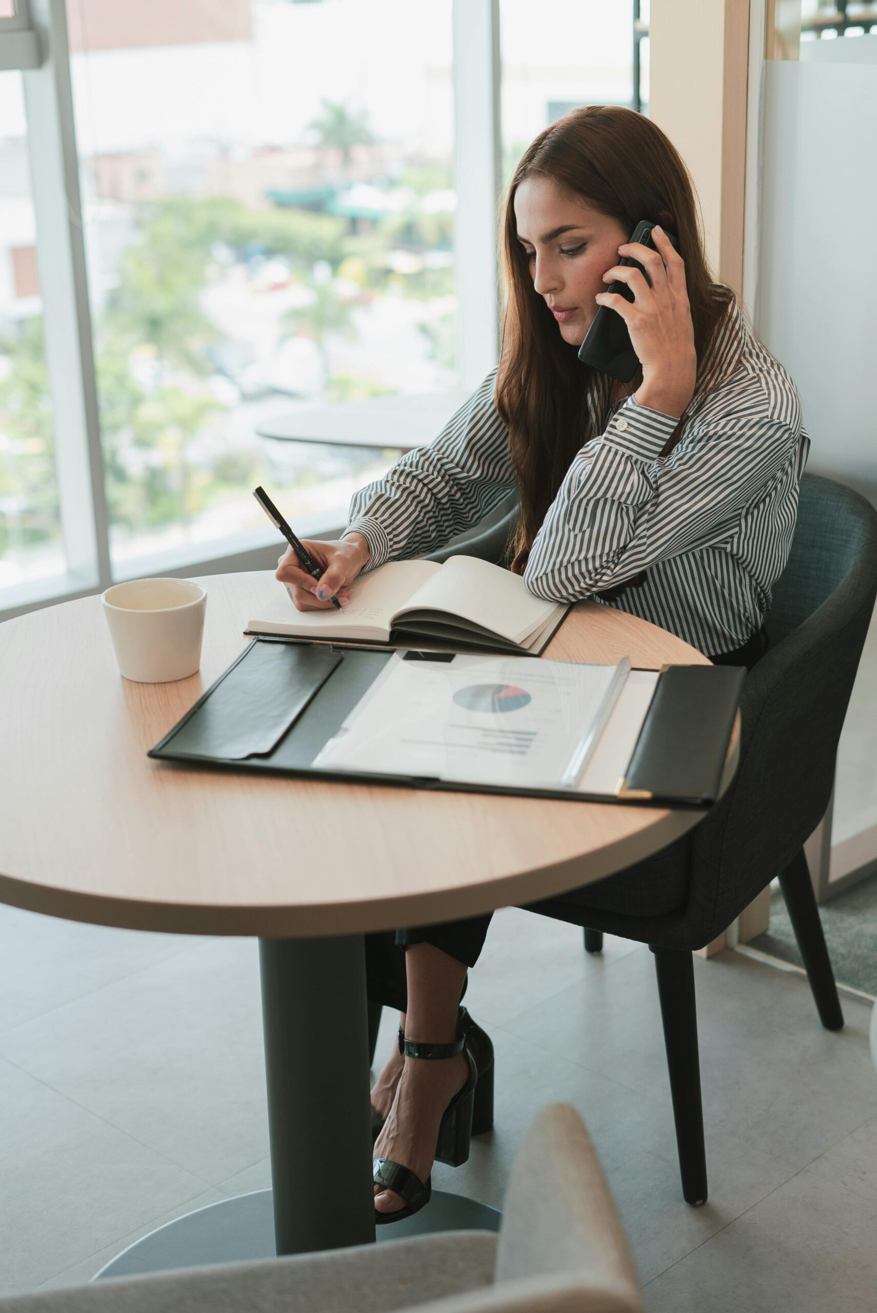 Woman in striped shirt multitasking in an office, writing notes while on a phone call, business setting.